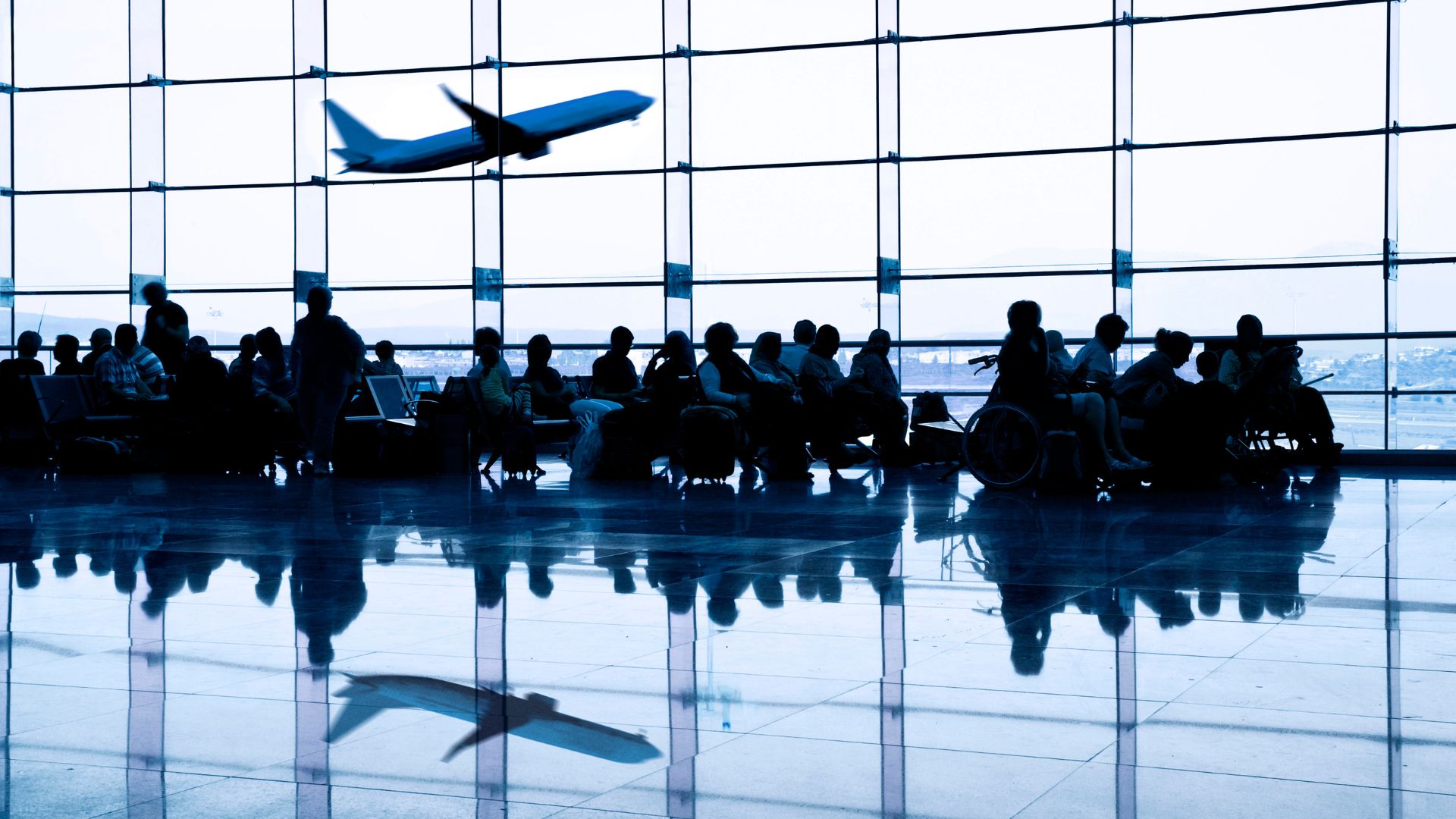 Passengers sitting at an airport terminal with a plane taking off in the background.